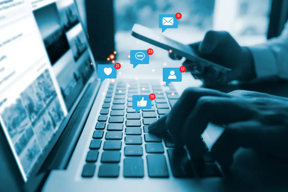 Hands using a laptop and smartphone with social media notification icons floating above.